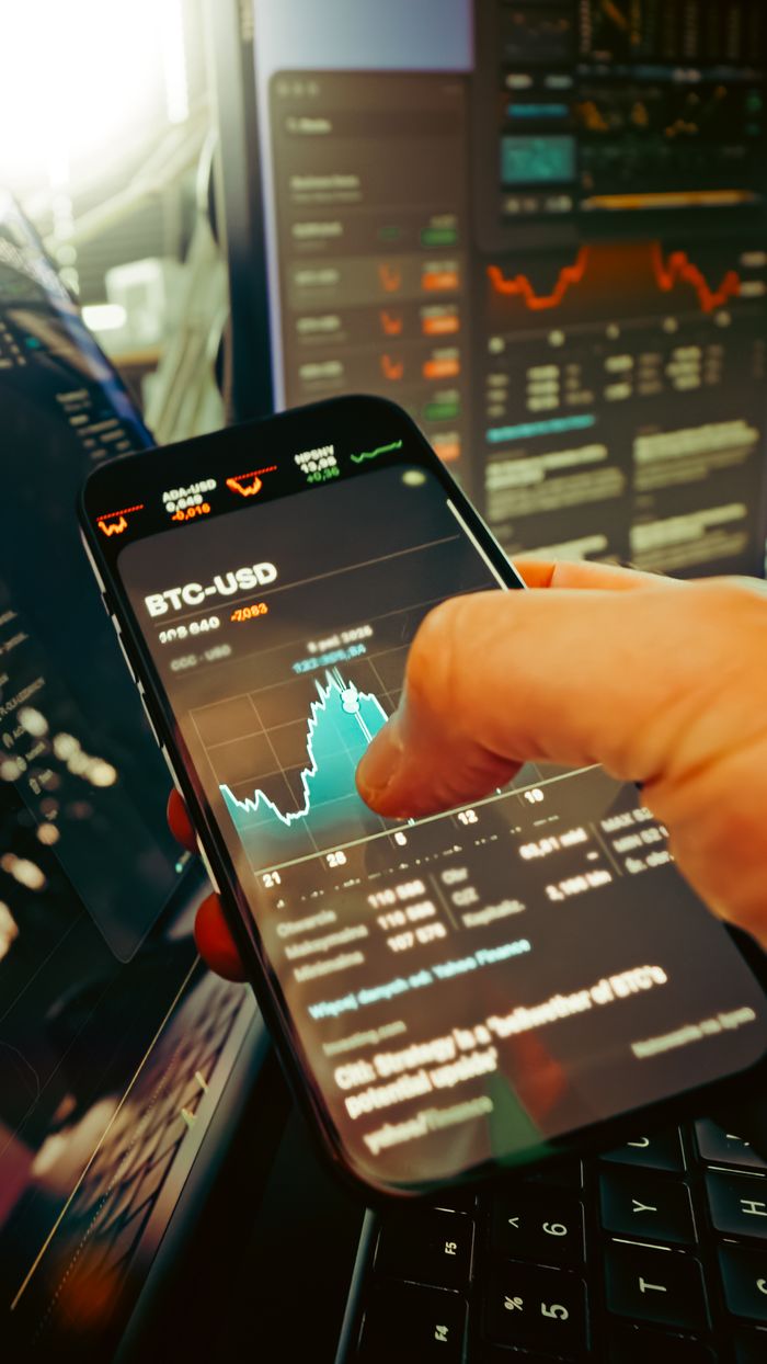Services A hand holds a smartphone displaying BTC-USD chart amidst digital trading screens, highlighting crypto finance.